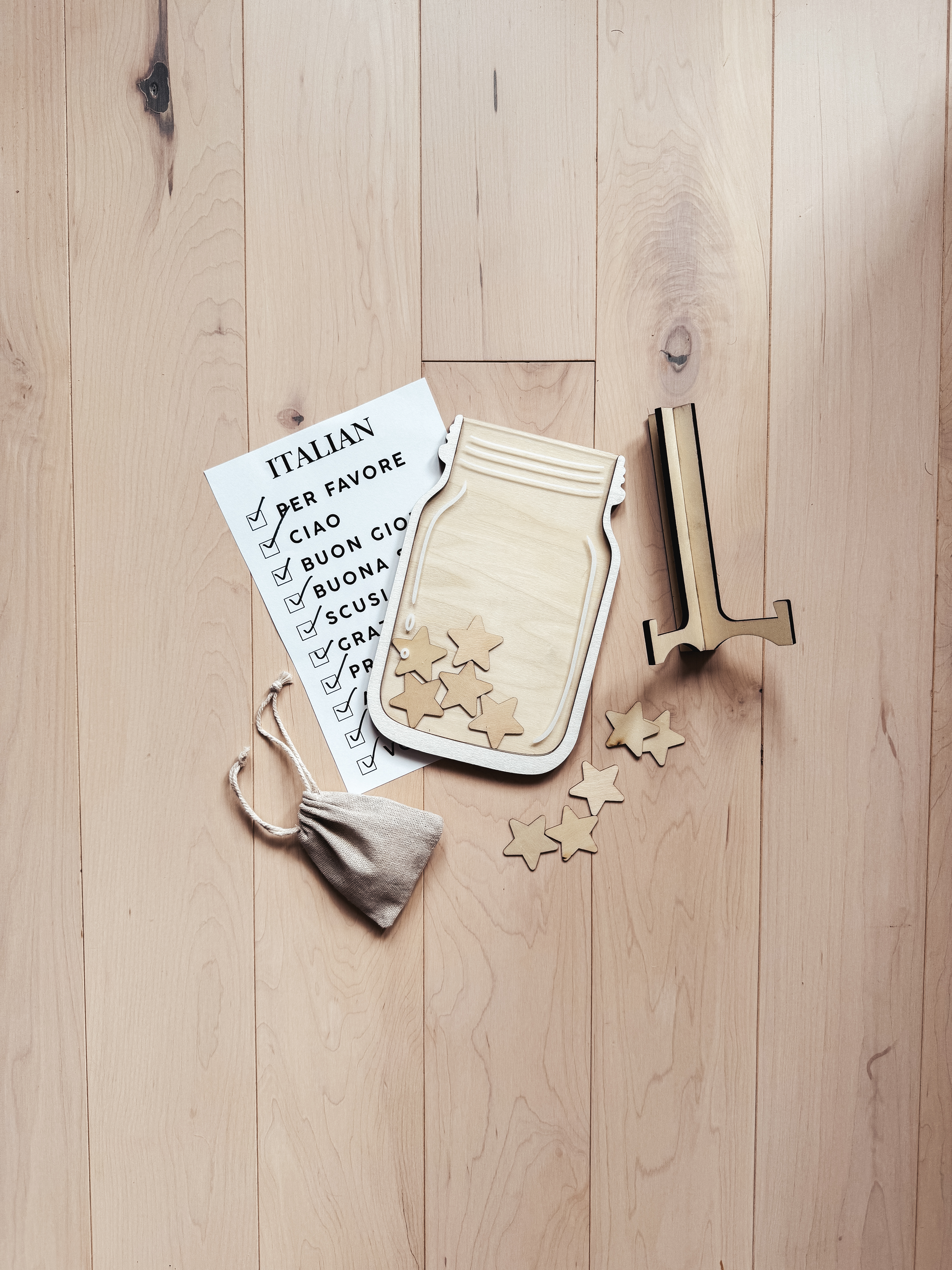 Wooden reward jar with star-shaped tokens and a checklist of basic Italian words like 'Ciao' and 'Per favore' for teaching kids a new language, accompanied by a fabric pouch for storing tokens.